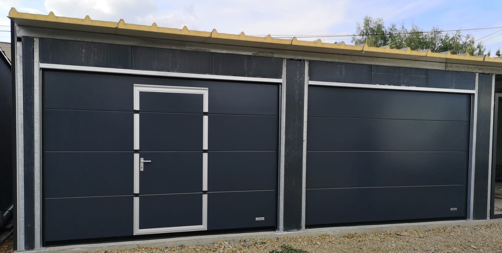 Szara brama garażowa segmentowa z drzwiami wejściowymi, widok z zewnątrz, jasne obramowanie, szara ściana budynku, żwirowe podłoże.