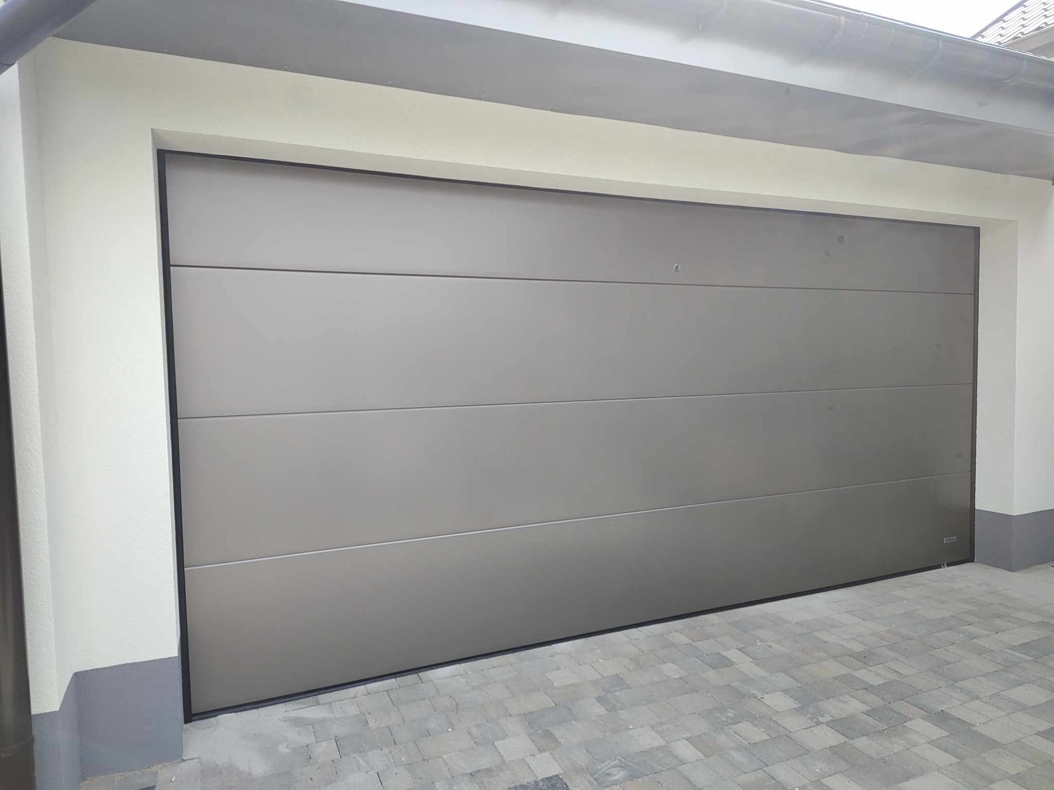 Zamknięta, segmentowa brama garażowa w kolorze szarym, z poziomymi przetłoczeniami, zamontowana w garażu w Rybniku. Widoczny fragment elewacji budynku i kostka brukowa przed garażem.