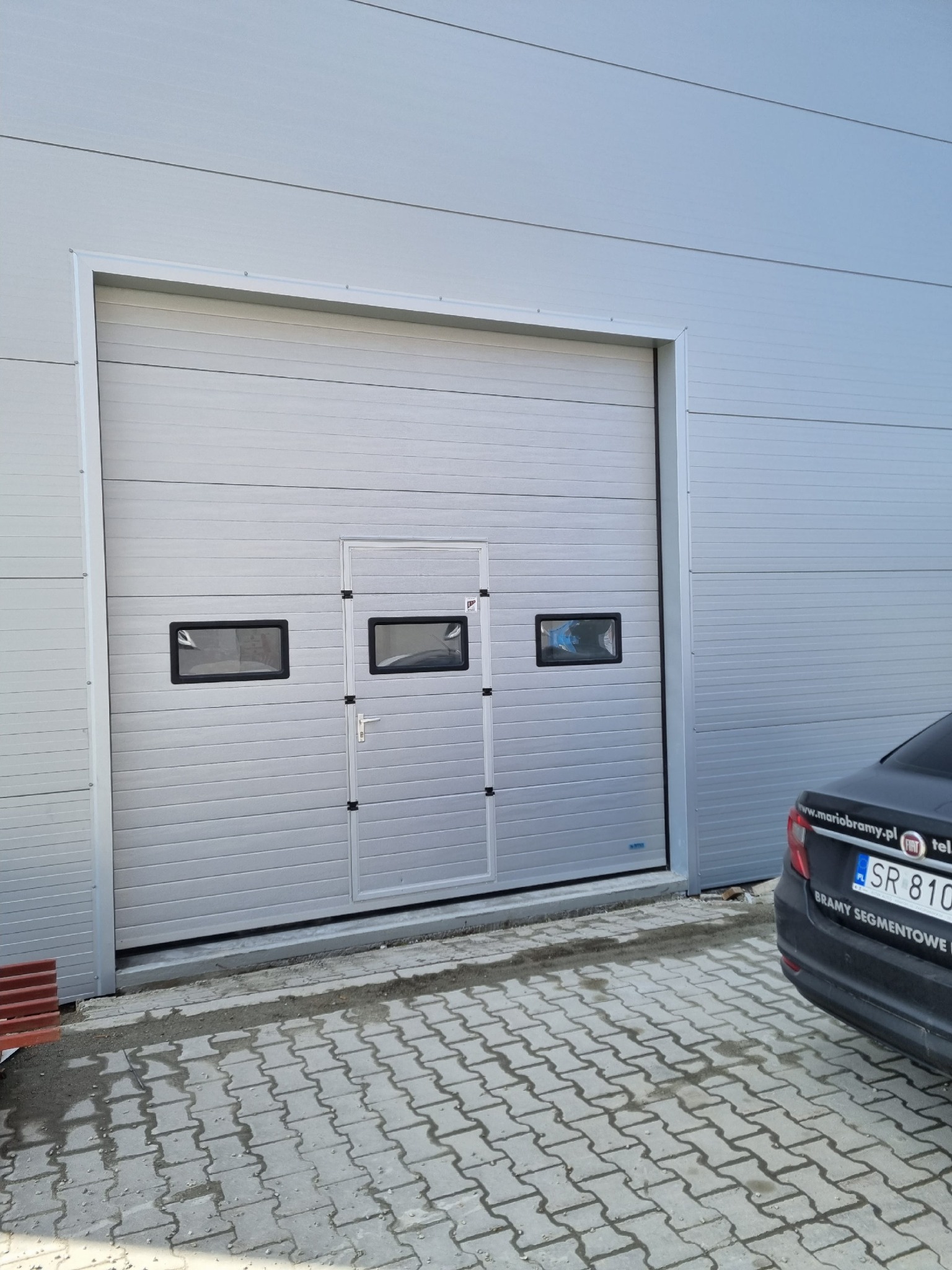 Szara segmentowa brama z oknami i drzwiami wejściowymi w budynku przemysłowym, widoczny fragment zaparkowanego samochodu osobowego.