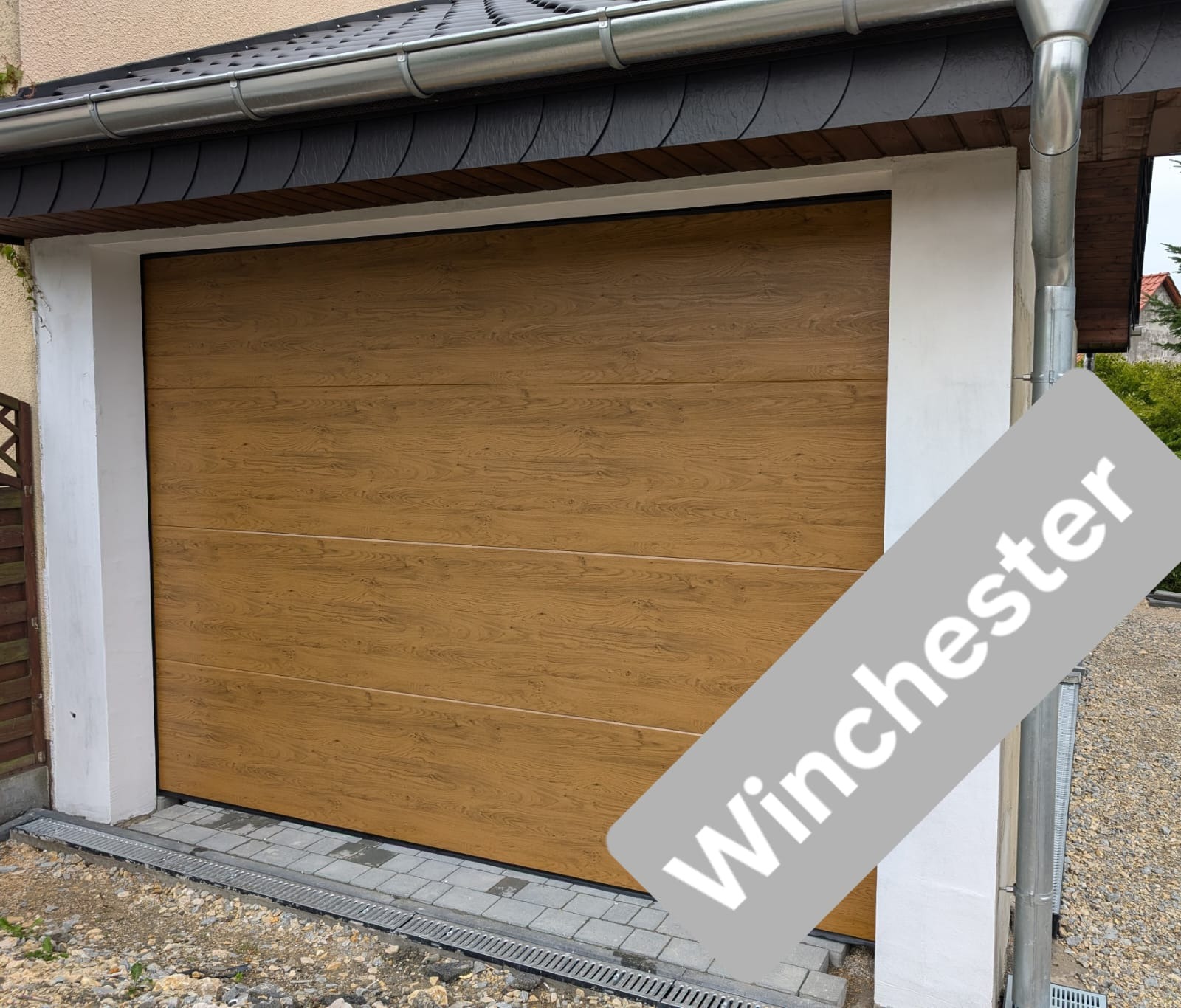 Zewnętrzny widok zamkniętej, segmentowej bramy garażowej w kolorze jasnego drewna, z widoczną fakturą usłojenia, zamontowanej w białym, otynkowanym garażu z szarą dachówką.