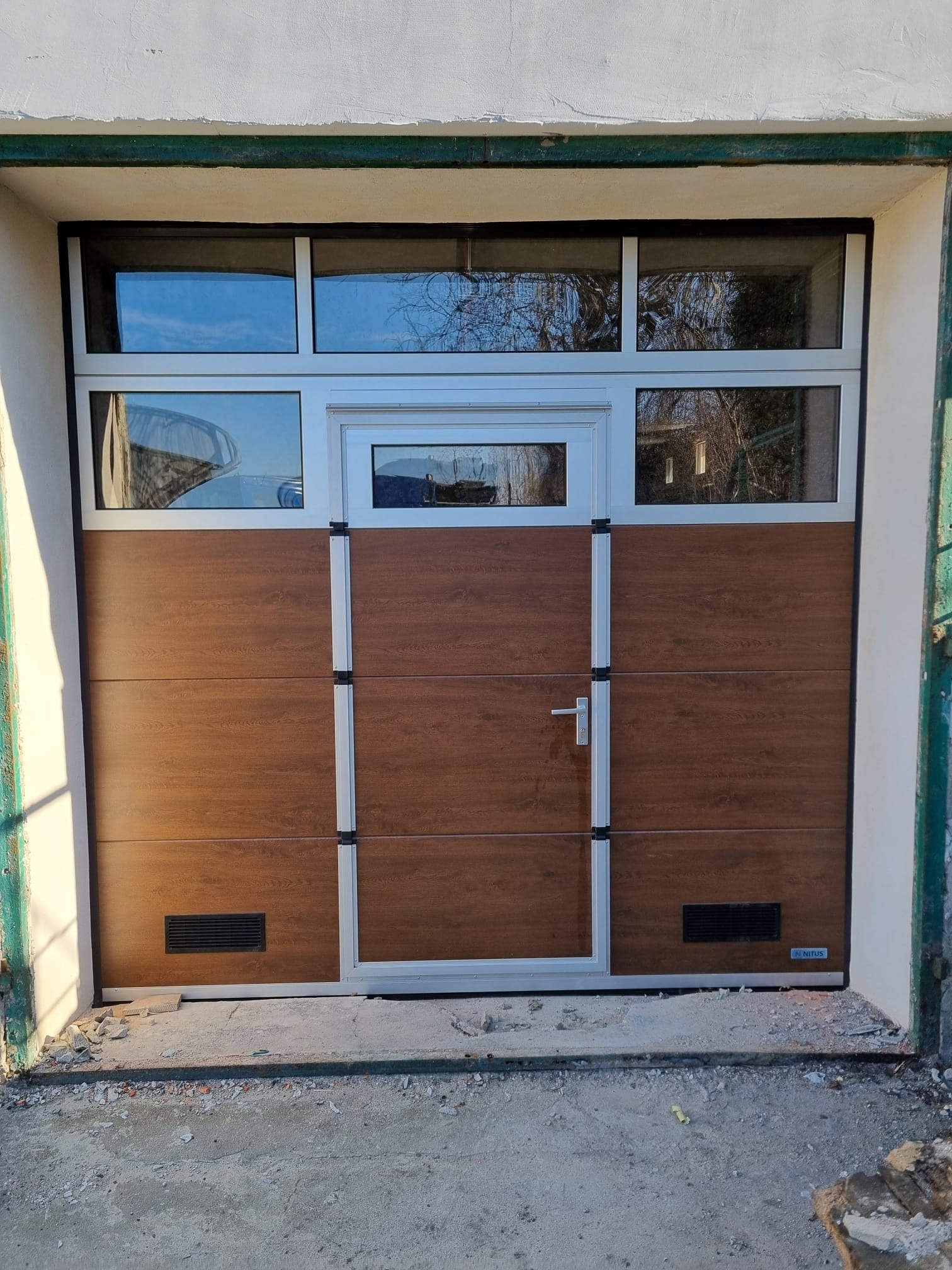 Nowa brama garażowa segmentowa z oknami i drzwiami wejściowymi, wykończona w kolorze drewna, widoczne niedokończone prace wykończeniowe wokół