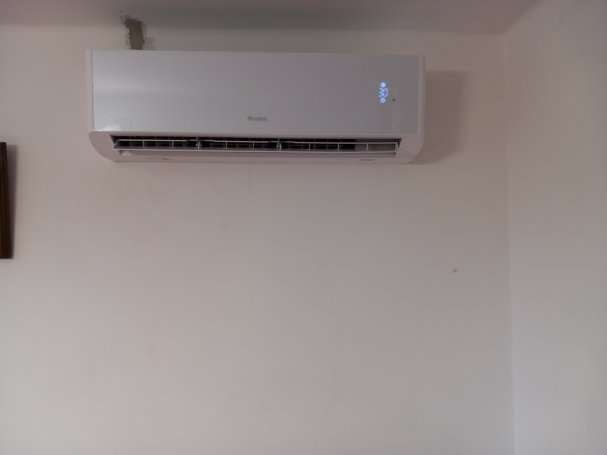 Biały klimatyzator marki Gree zamontowany na ścianie, wyświetlający temperaturę 30 stopni, widoczne ślady po naprawach na ścianie nad urządzeniem.
