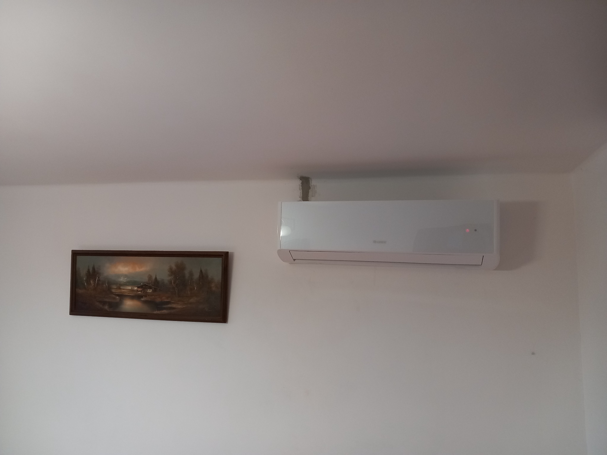 Biały klimatyzator ścienny zamontowany nad obrazem z pejzażem w brązowej ramie, widoczne szare rury przy suficie.