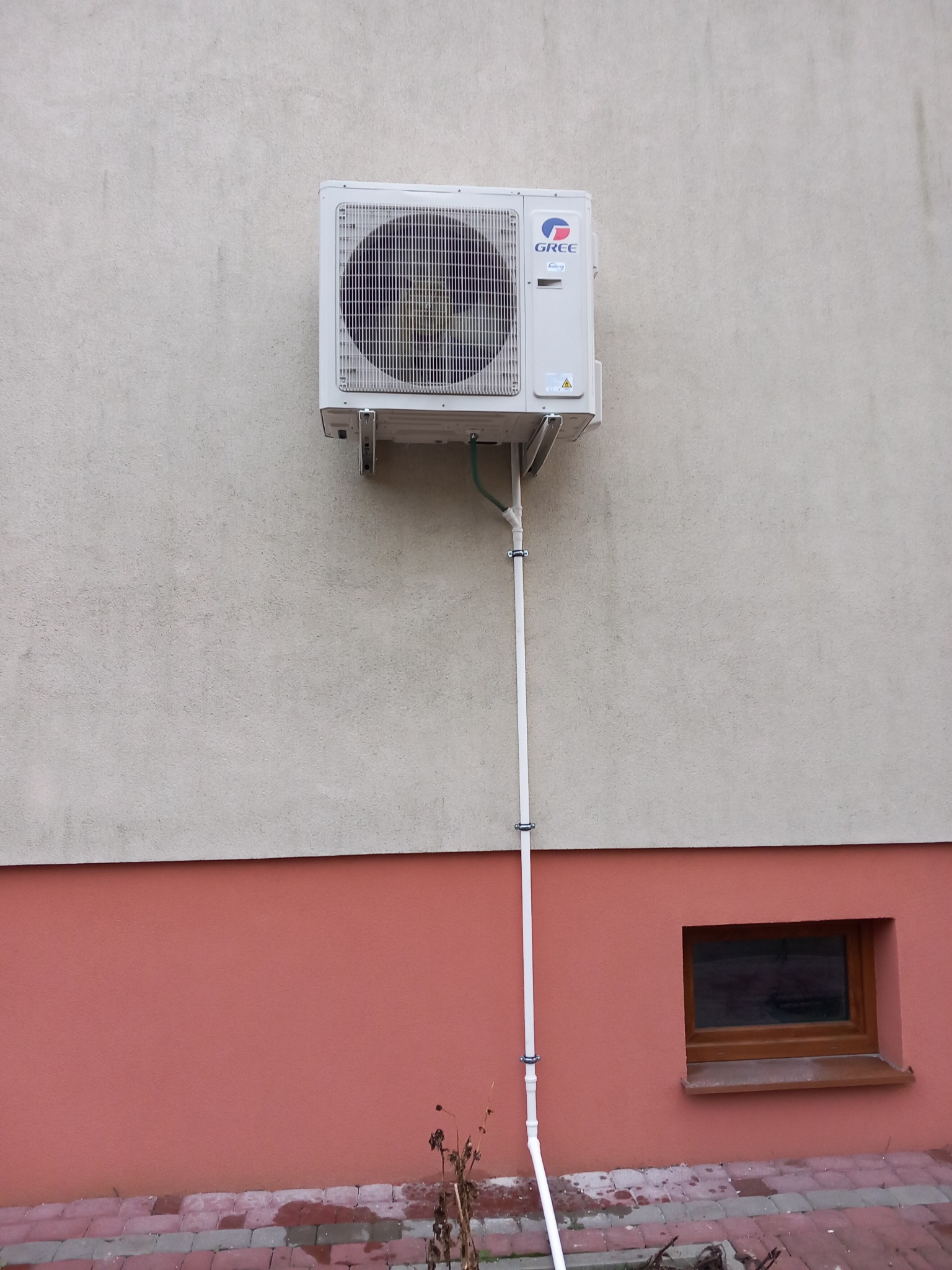 Zewnętrzna jednostka klimatyzacji marki Gree zamontowana na ścianie budynku z widocznymi rurami odprowadzającymi skropliny.