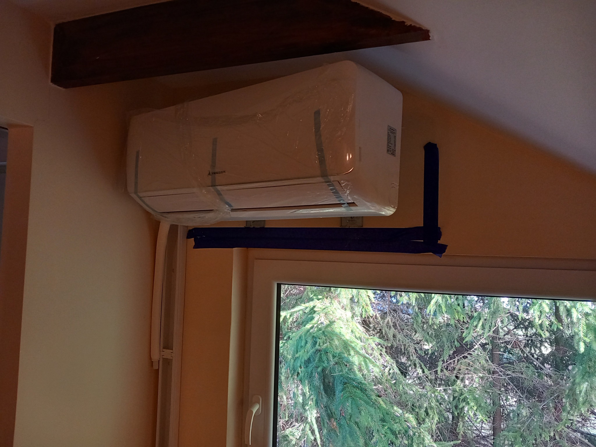 Biała jednostka klimatyzacji Mitsubishi, owinięta folią ochronną, zamontowana na ścianie nad oknem, zabezpieczona taśmą malarską, z widocznym fragmentem drewnianej belki stropowej.