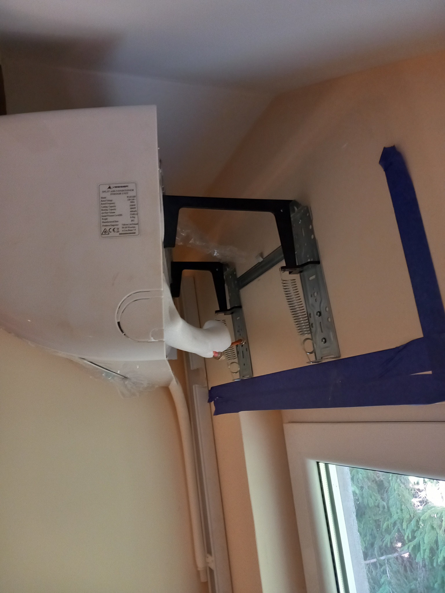 Wewnętrzna jednostka klimatyzacji marki Mitsubishi, zamontowana na metalowym wsporniku przy ścianie, zabezpieczona niebieską taśmą malarską podczas instalacji, widoczne rury w izolacji oraz okno...