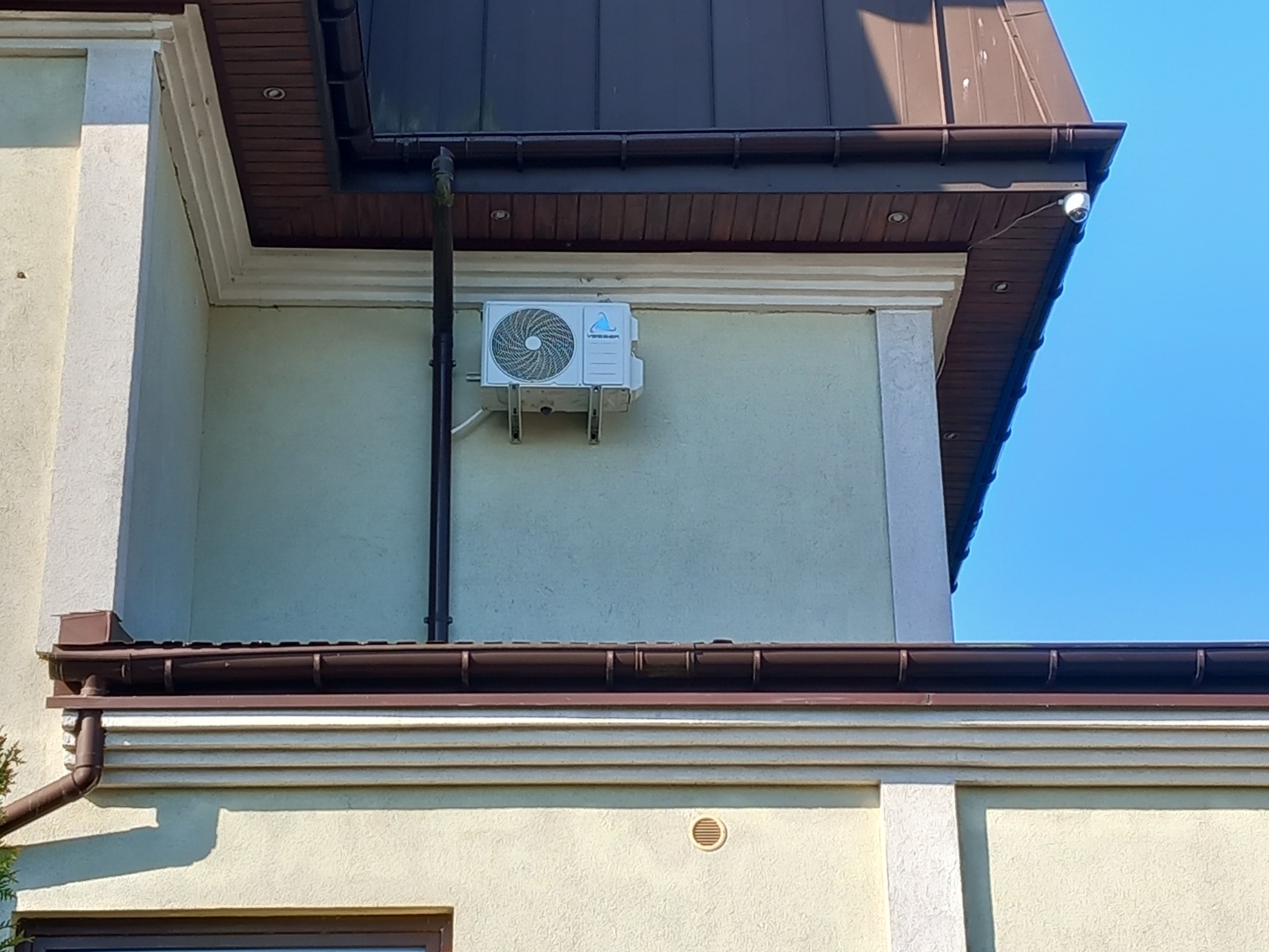 Zewnętrzna jednostka klimatyzatora zamontowana na elewacji budynku z brązowymi rynnami i dachem, widoczna kamera monitoringu.