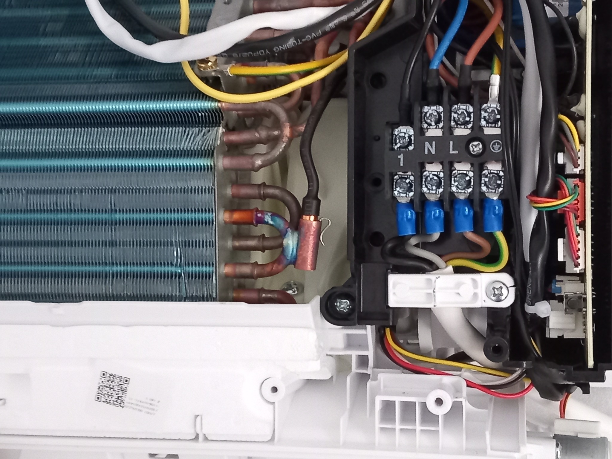 Wnętrze klimatyzatora z widocznymi miedzianymi rurkami, żebrowanym wymiennikiem ciepła, kostką elektryczną z przewodami o różnych kolorach oraz kodem QR na białej obudowie.