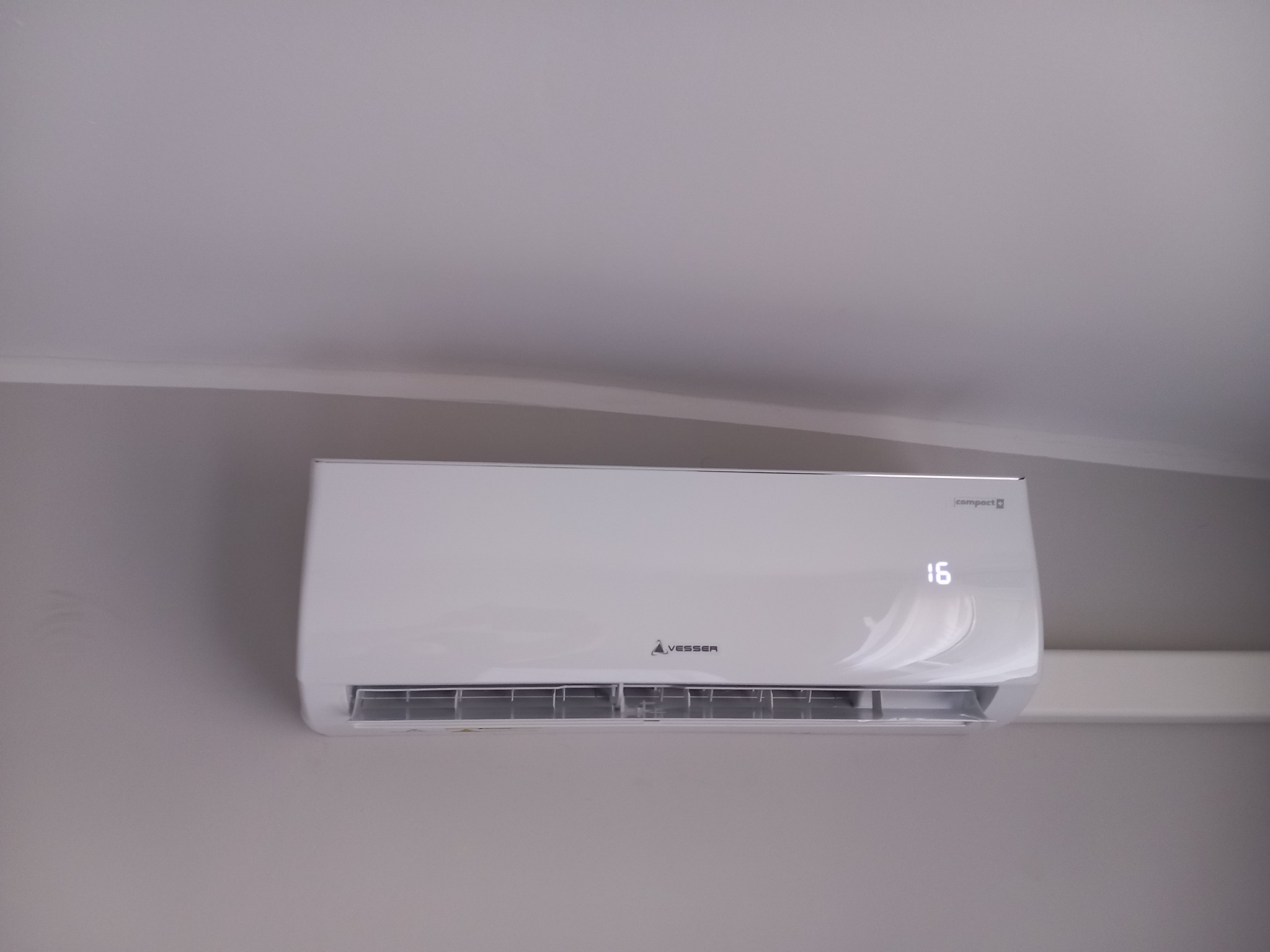 Biały klimatyzator Vesser Compact 16 zamontowany na szarej ścianie, widoczny wyświetlacz temperatury.