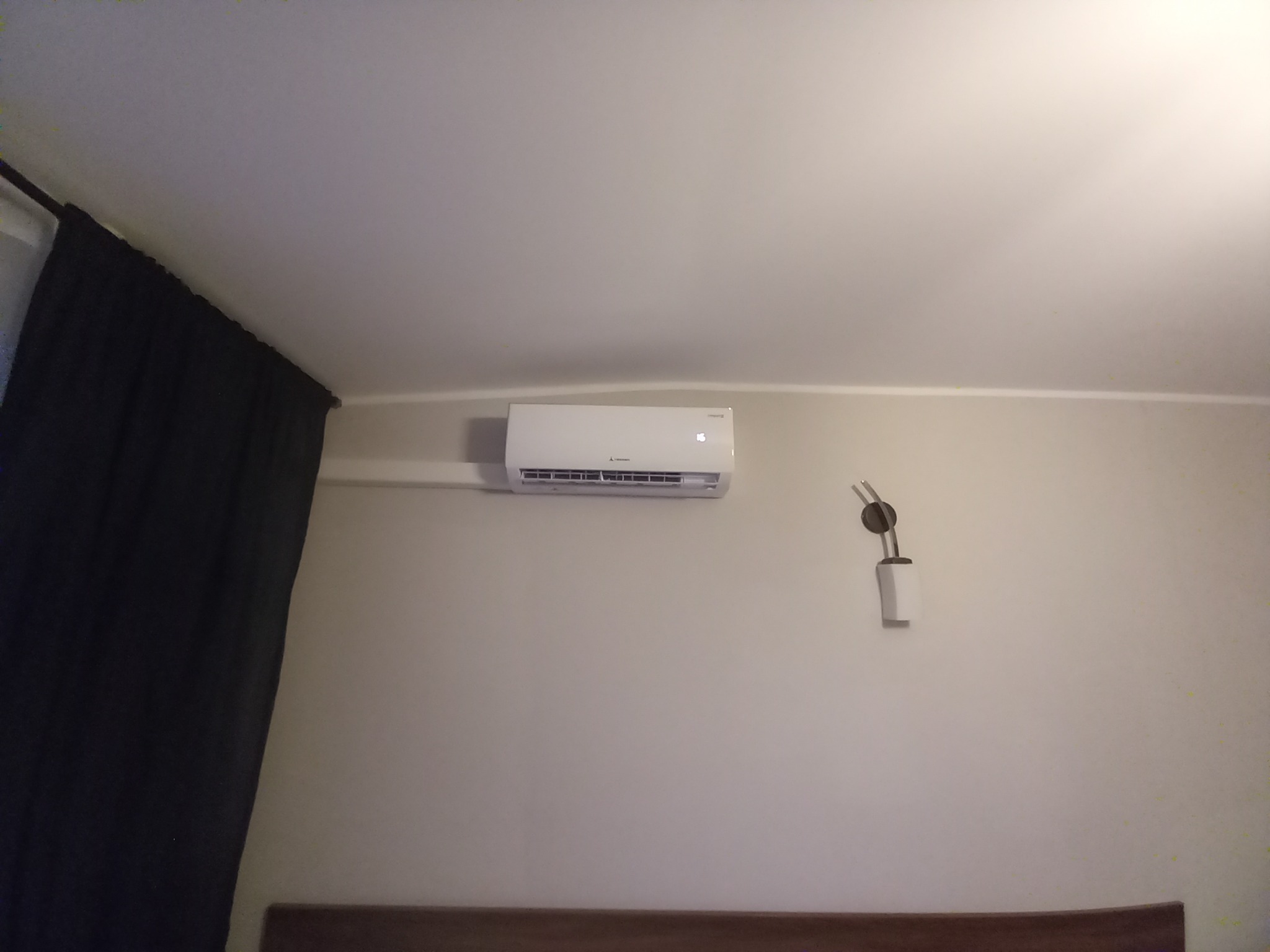 Biały klimatyzator Mitsubishi Electric zamontowany na ścianie w pokoju z beżowymi ścianami, widoczna lampka ścienna i fragment ciemnych zasłon.