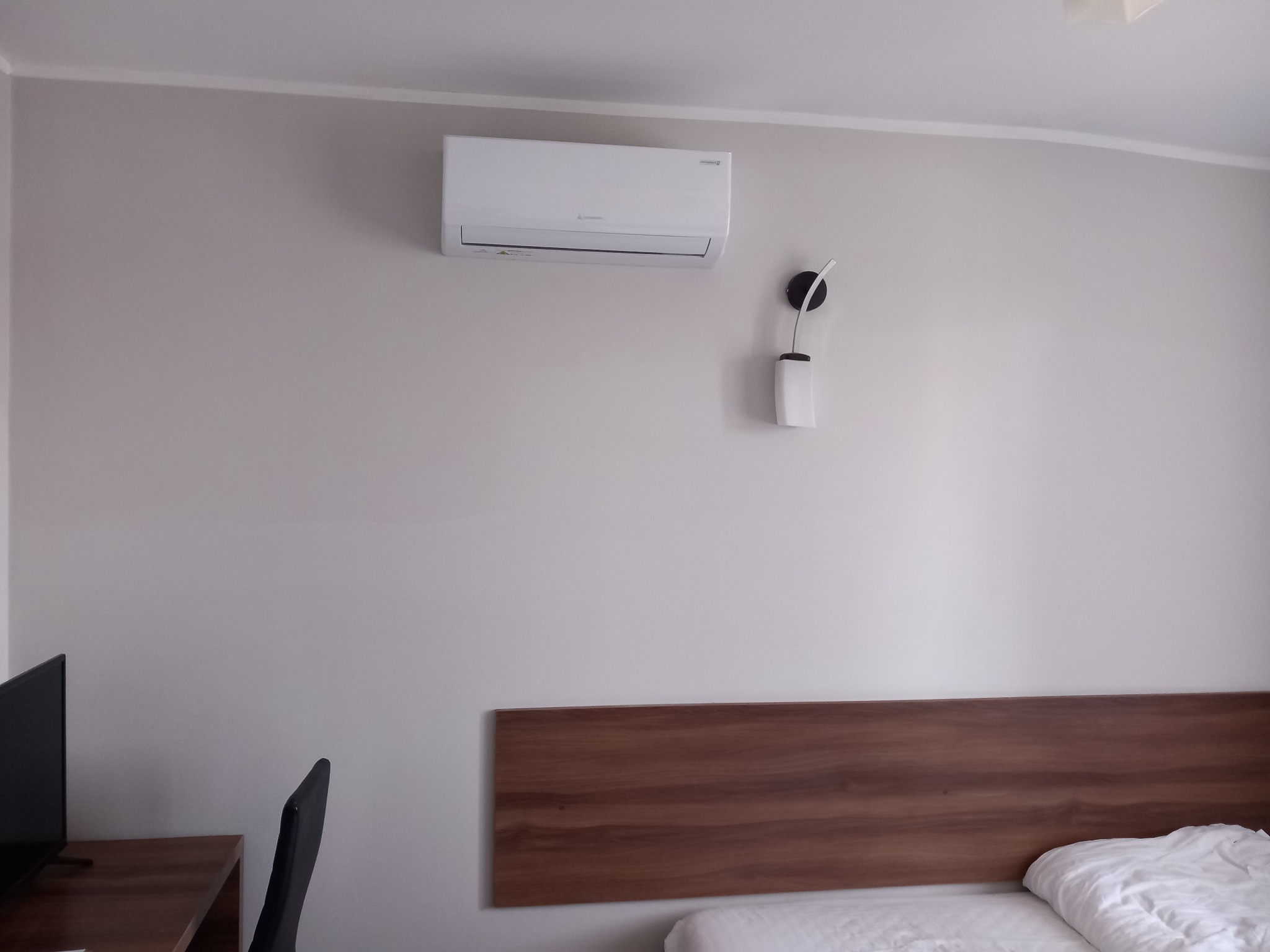Biały klimatyzator ścienny zamontowany nad drewnianym zagłówkiem łóżka w pokoju hotelowym, obok lampka nocna.