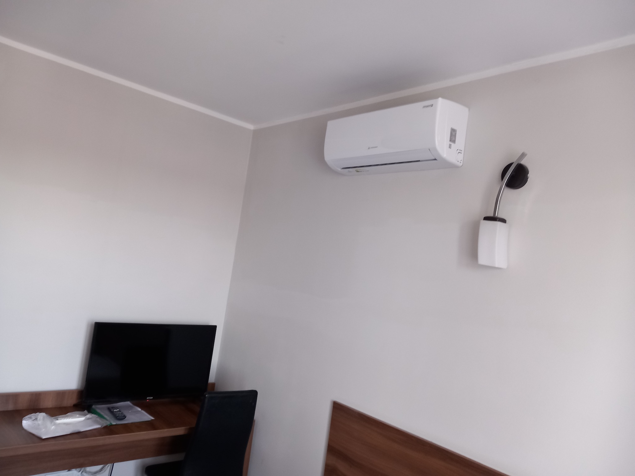 Biały klimatyzator ścienny Mitsubishi Electric w pokoju hotelowym, obok lampka nocna z regulowanym ramieniem, biurko z telewizorem i krzesłem.