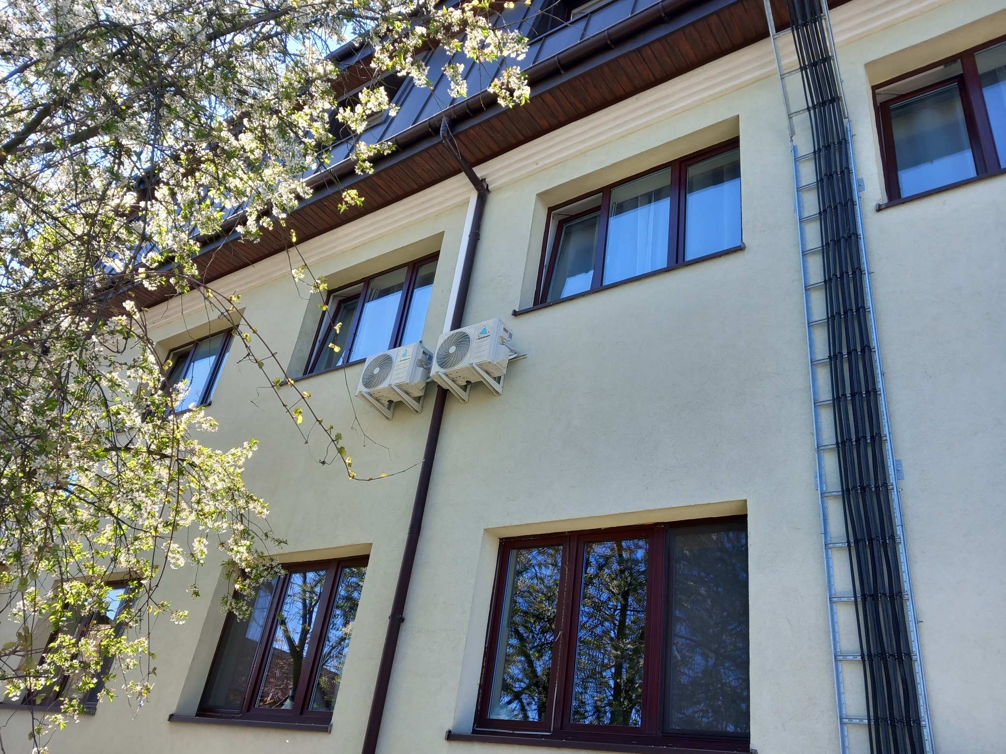 Zewnętrzny widok budynku w Warszawie z zamontowanymi dwoma klimatyzatorami na ścianie, widoczne okna z odbiciem drzew, rynna i drabinka kablowa, z kwitnącym drzewem w rogu kadru.