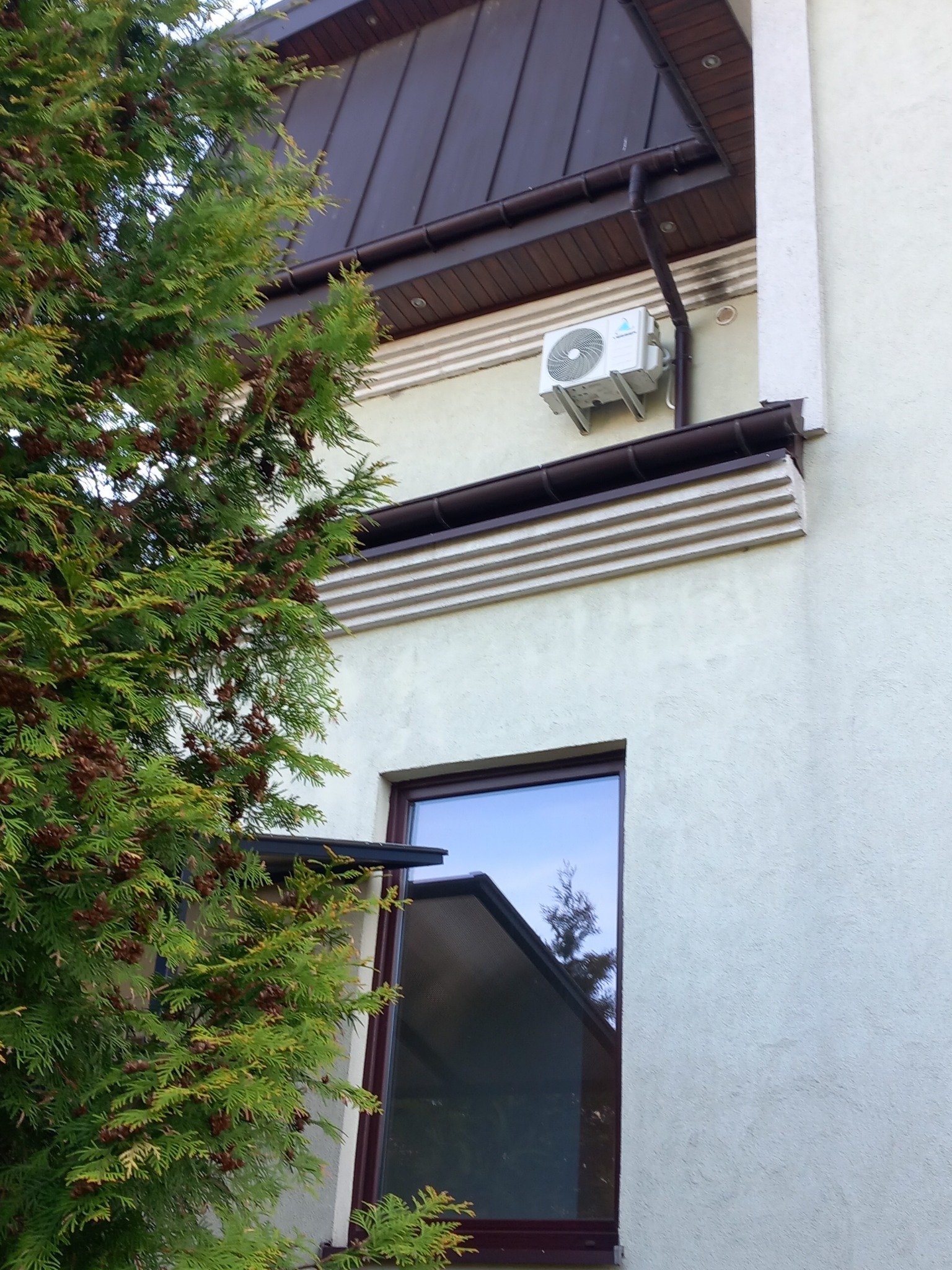 Zewnętrzna jednostka klimatyzacji zamontowana na elewacji domu, widoczna zza iglastego drzewa, z brązowym dachem i rynnami w tle.