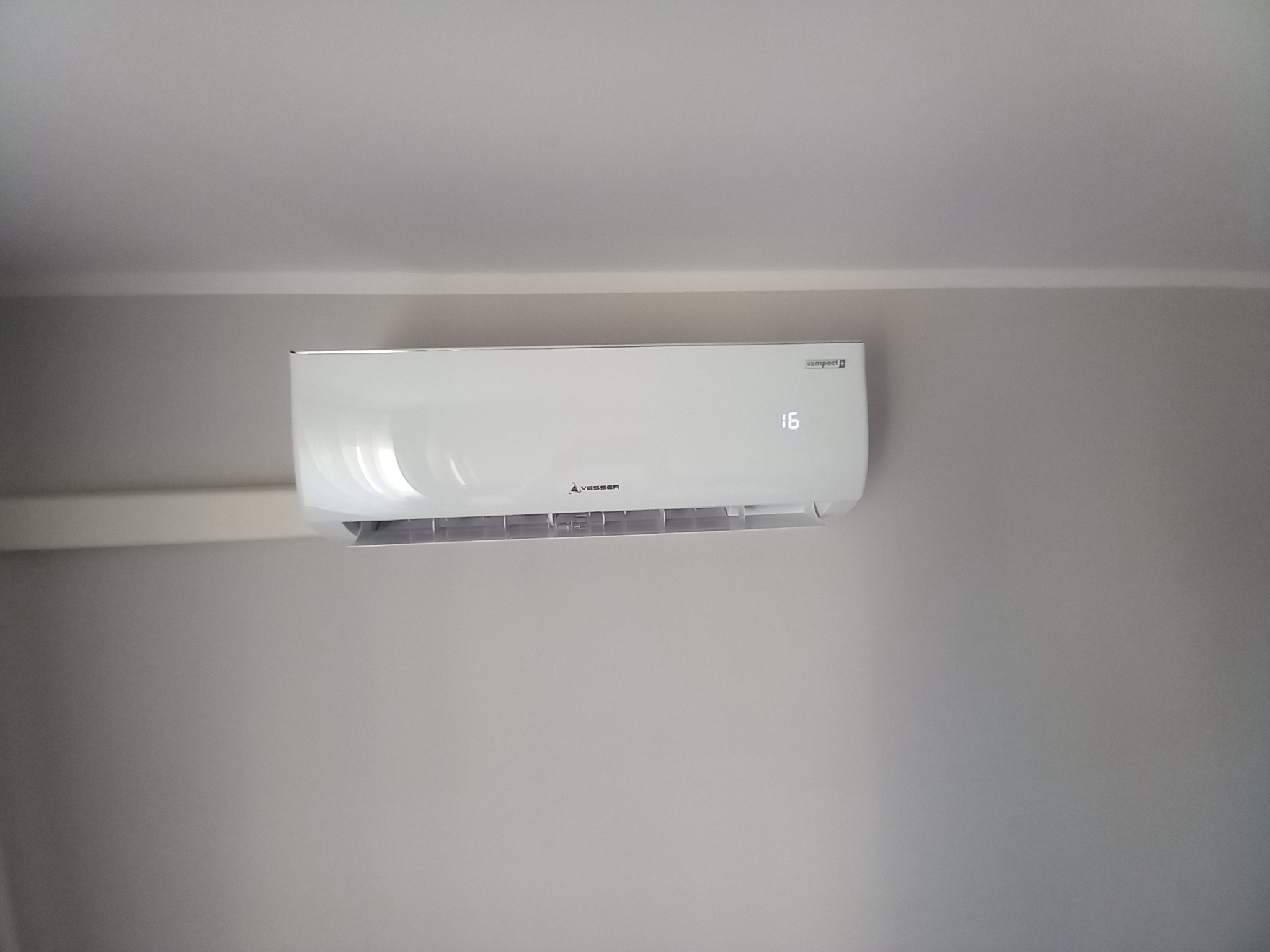 Biała jednostka klimatyzacyjna Vesser Compact+ zamontowana na szarej ścianie, wyświetlająca temperaturę 16 stopni.