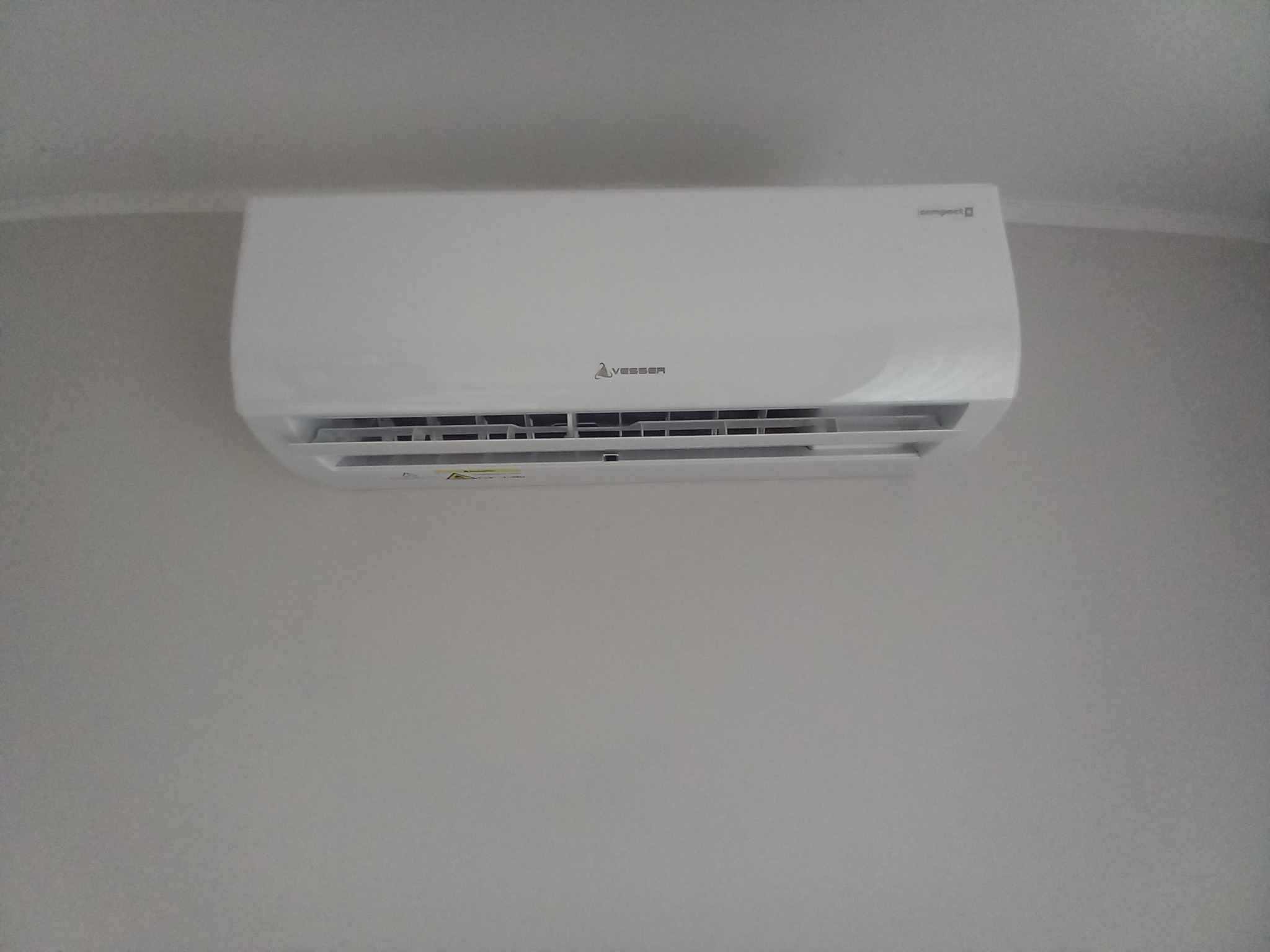 Biała jednostka klimatyzacyjna marki Vesser zamontowana na jasnej ścianie w pomieszczeniu. Widoczne logo i napisy na obudowie.