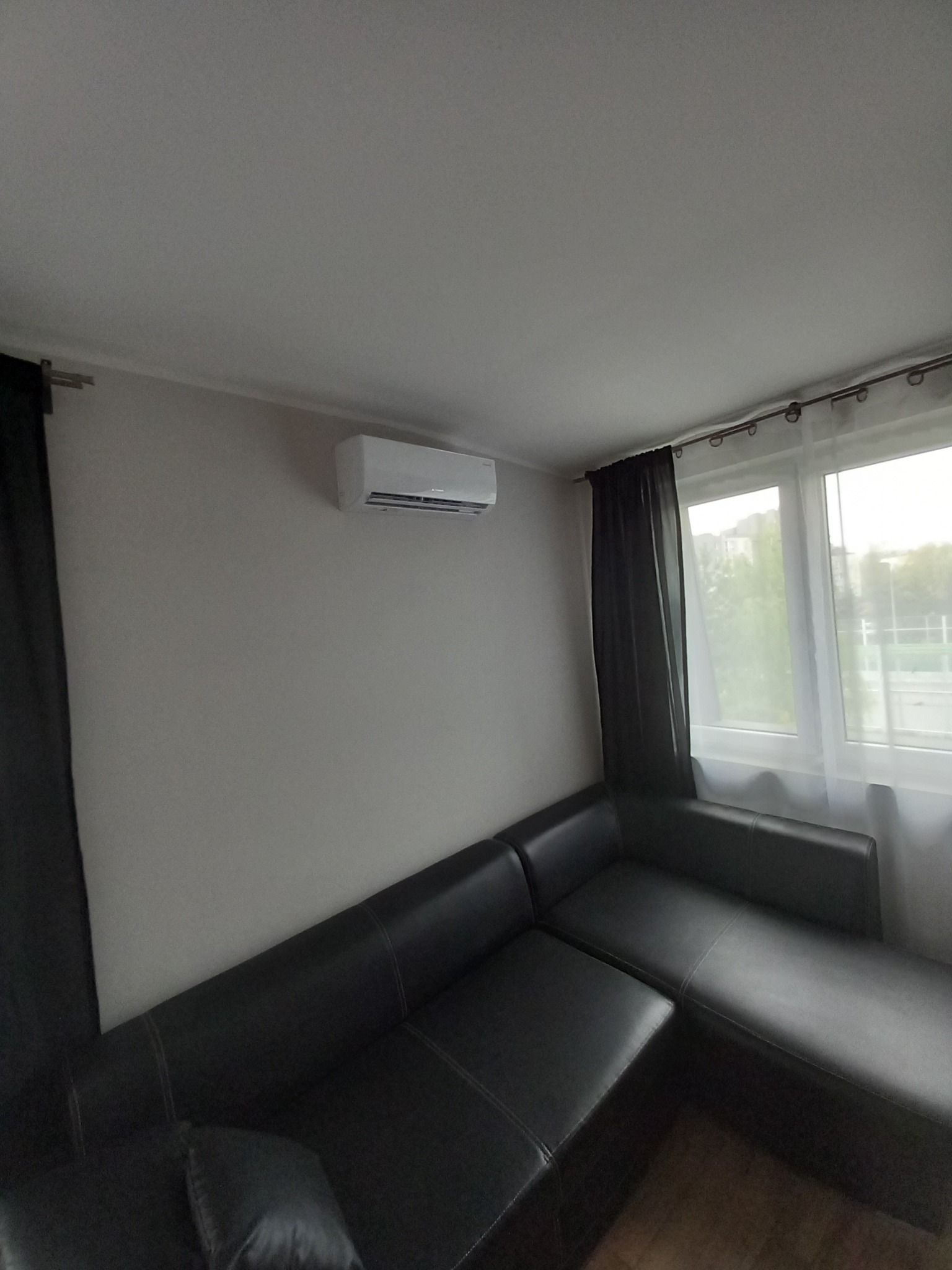 Biała jednostka klimatyzacyjna zamontowana na ścianie nad czarnym narożnikiem w pokoju z oknem zasłoniętym częściowo czarną i białą zasłoną.