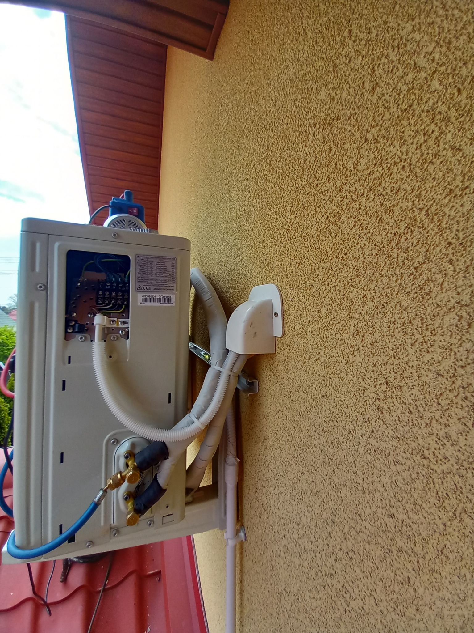 Zewnętrzna jednostka klimatyzacji w trakcie montażu na elewacji budynku, z otwartym panelem elektrycznym i podłączonymi przewodami chłodniczymi.