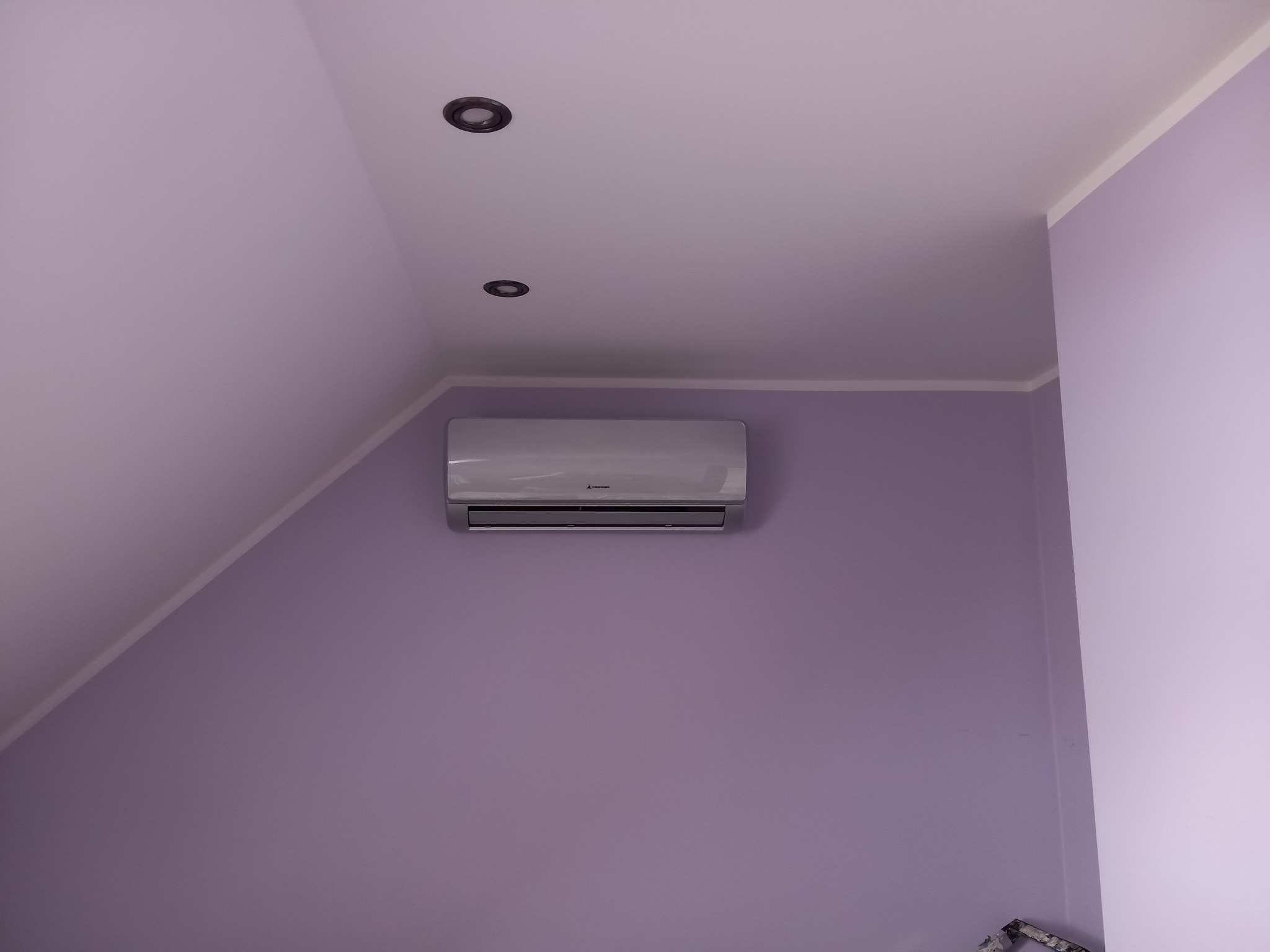 Biały klimatyzator Mitsubishi Electric zamontowany na fioletowej ścianie pod skosem, z widocznymi dwoma punktowymi lampami sufitowymi.