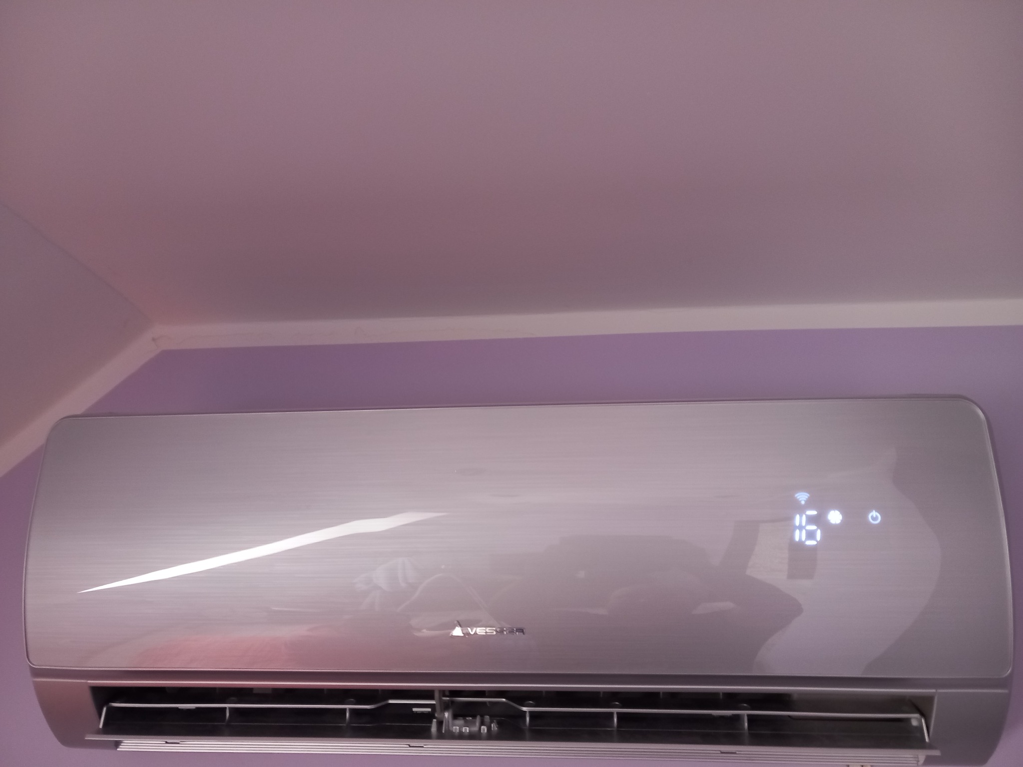 Zamontowany srebrny klimatyzator z wyświetlaczem temperatury 16 stopni Celsjusza na fioletowej ścianie w pomieszczeniu.