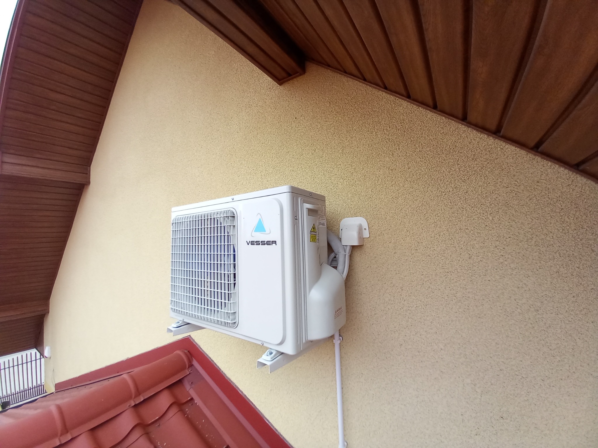 Biała jednostka zewnętrzna klimatyzacji marki Vesser zamontowana na beżowej elewacji domu, widoczna instalacja elektryczna i rury chłodnicze, fragment dachu z czerwonej dachówki w dolnej części kadru.