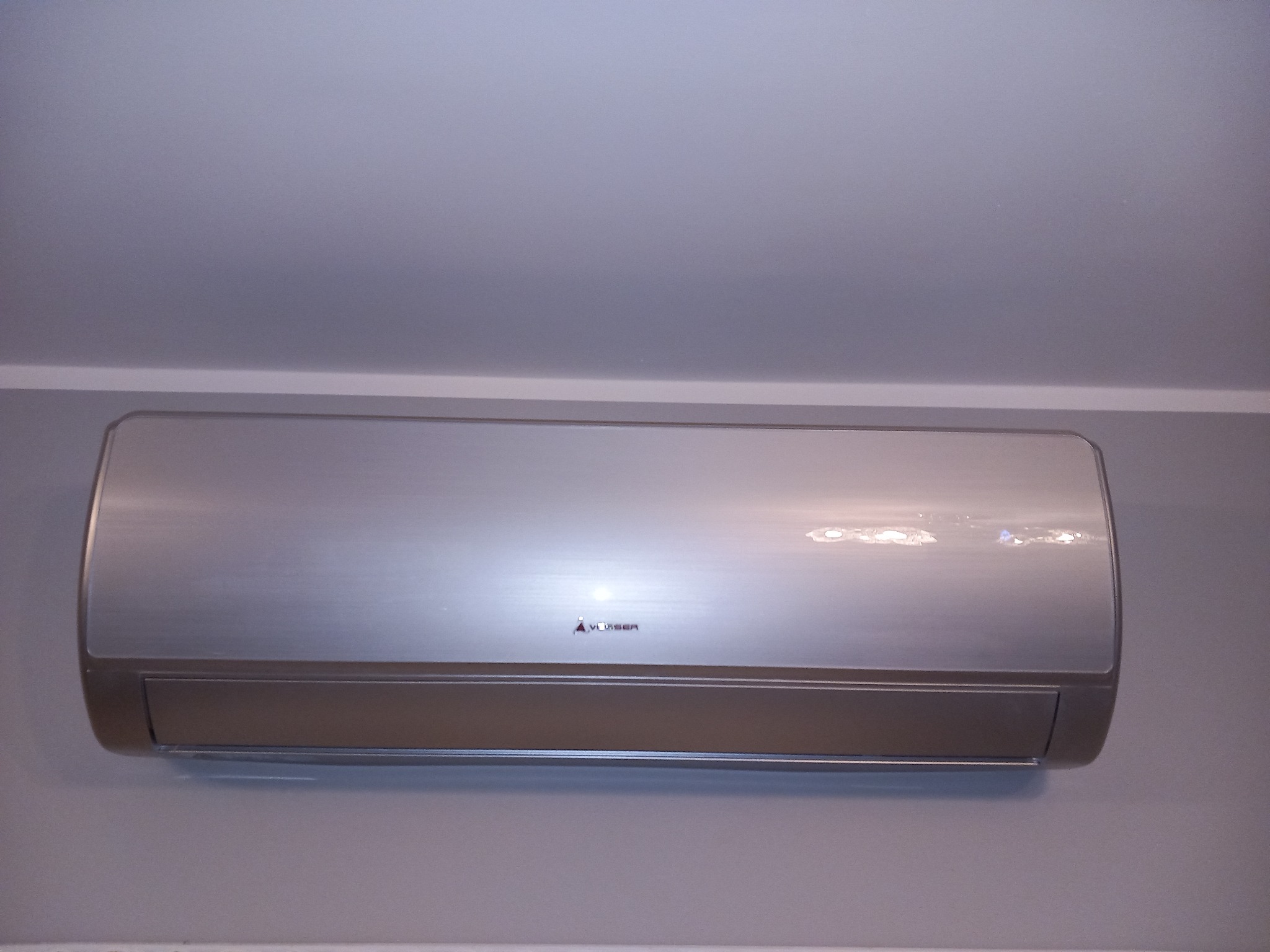 Zamontowany srebrny klimatyzator ścienny marki Vesser na szarej ścianie, widok z dołu.