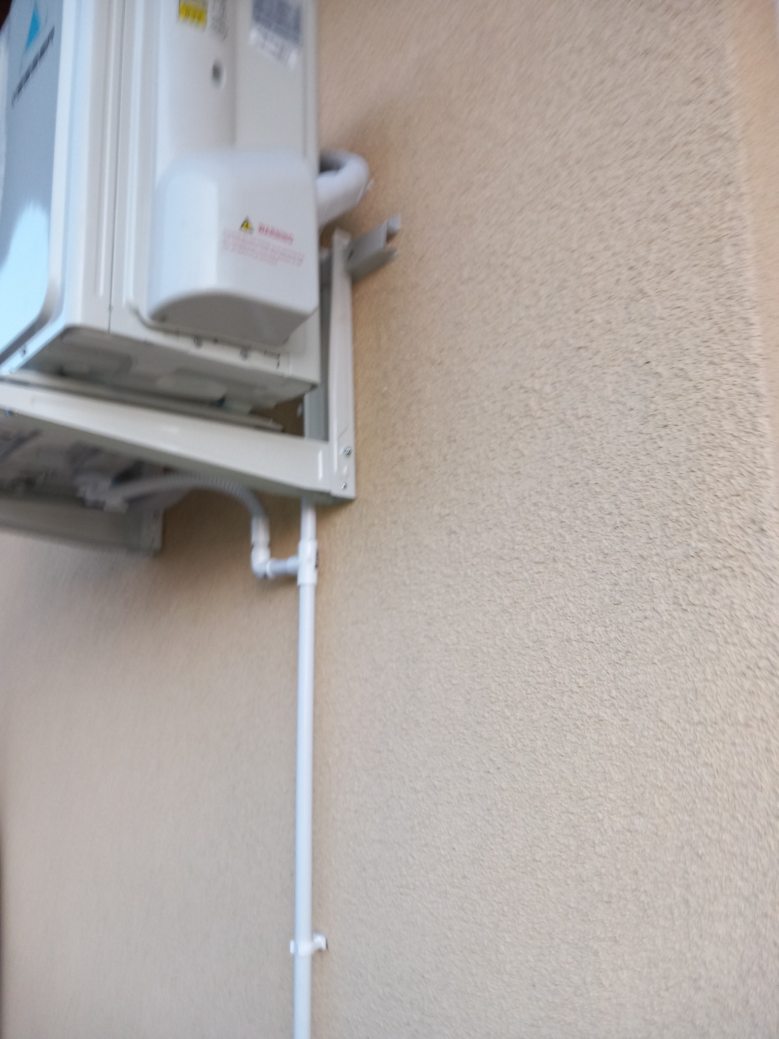 Zewnętrzna jednostka klimatyzacji zamontowana na beżowej, teksturowanej ścianie budynku z widocznymi rurami odprowadzającymi skropliny.