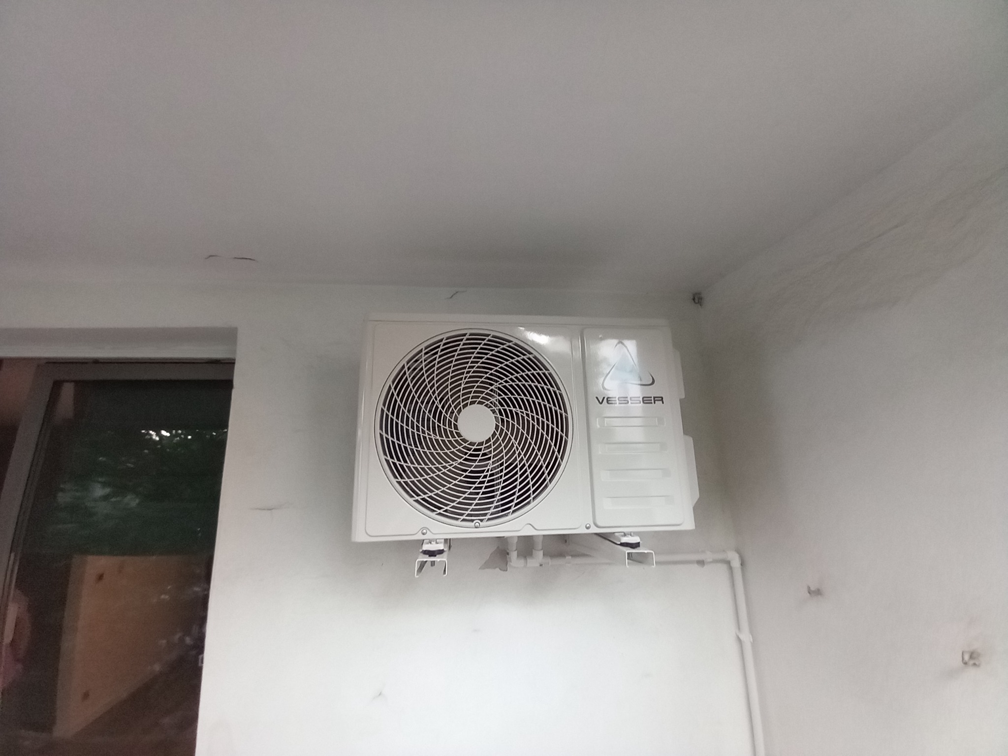 Biała jednostka wewnętrzna klimatyzacji marki Vesser zamontowana na ścianie w pomieszczeniu z widoczną instalacją rur. Ściana wymaga odświeżenia.