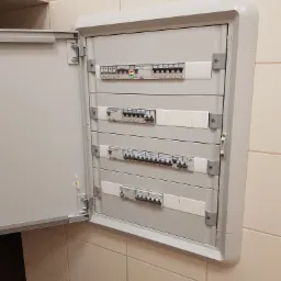 Otwarta szara skrzynka z bezpiecznikami i wyłącznikami różnicowoprądowymi zamontowana na kafelkowanej ścianie.