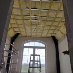 Poddasze w trakcie ocieplania wełną mineralną, widoczne rusztowanie drewniane i drabina, okno łukowe w tle.