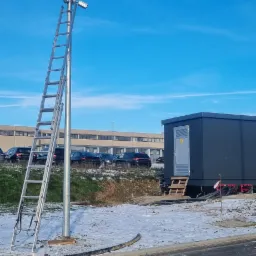 Metalowa latarnia z zamontowanymi kamerami monitoringu, przystawiona drabina, w tle budynek i zaparkowane samochody, częściowo ośnieżony teren.