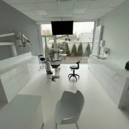 Jasne wnętrze gabinetu z fotelem dentystycznym, mikroskopem na ścianie i widokiem na ogród przez okno.