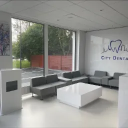 Jasne, minimalistyczne wnętrze poczekalni kliniki z szarymi sofami, białym stolikiem i recepcją, widoczne logo 'City Dental' na ścianie, obraz abstrakcyjny na ścianie po lewej stronie, duże okna...