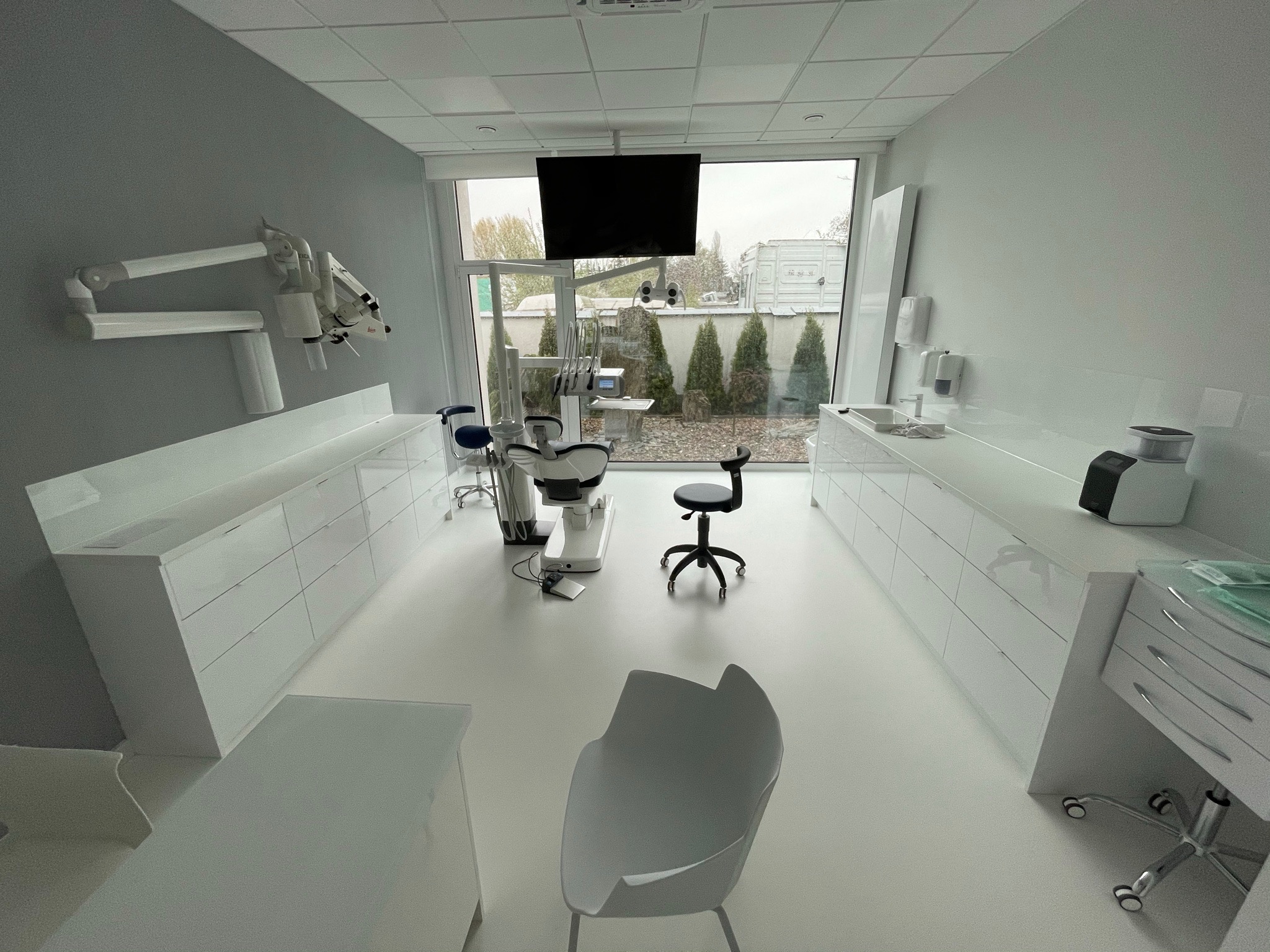 Jasne wnętrze gabinetu z fotelem dentystycznym, mikroskopem na ścianie i widokiem na ogród przez okno.