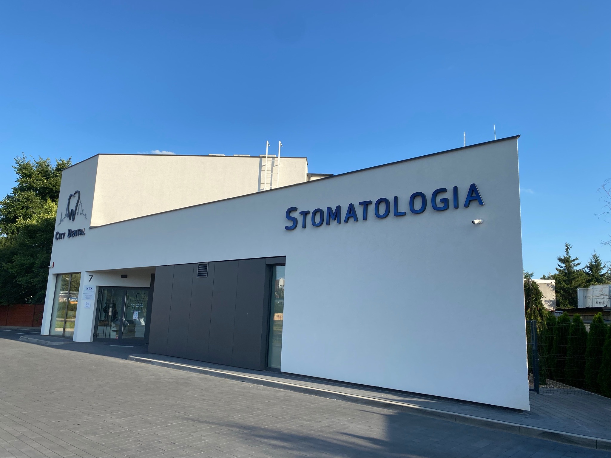 Nowoczesny budynek kliniki z napisem 'Stomatologia' w niebieskim kolorze na białej fasadzie, widok z zewnątrz w słoneczny dzień.