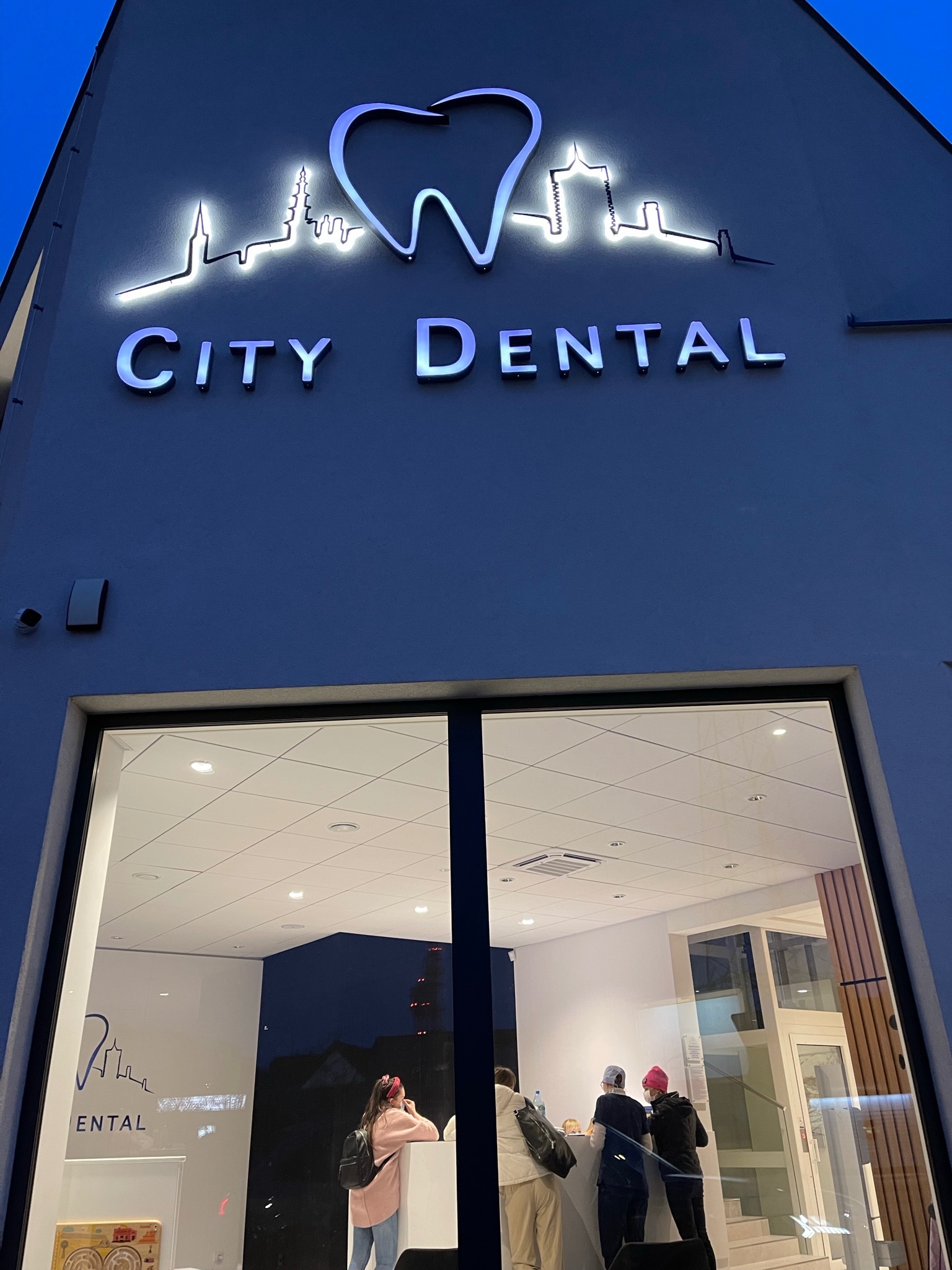Elewacja budynku z podświetlanym logo dentystycznym przedstawiającym zarys zęba i panoramy miasta nad napisem 'City Dental', widoczne osoby w recepcji przez duże okno.