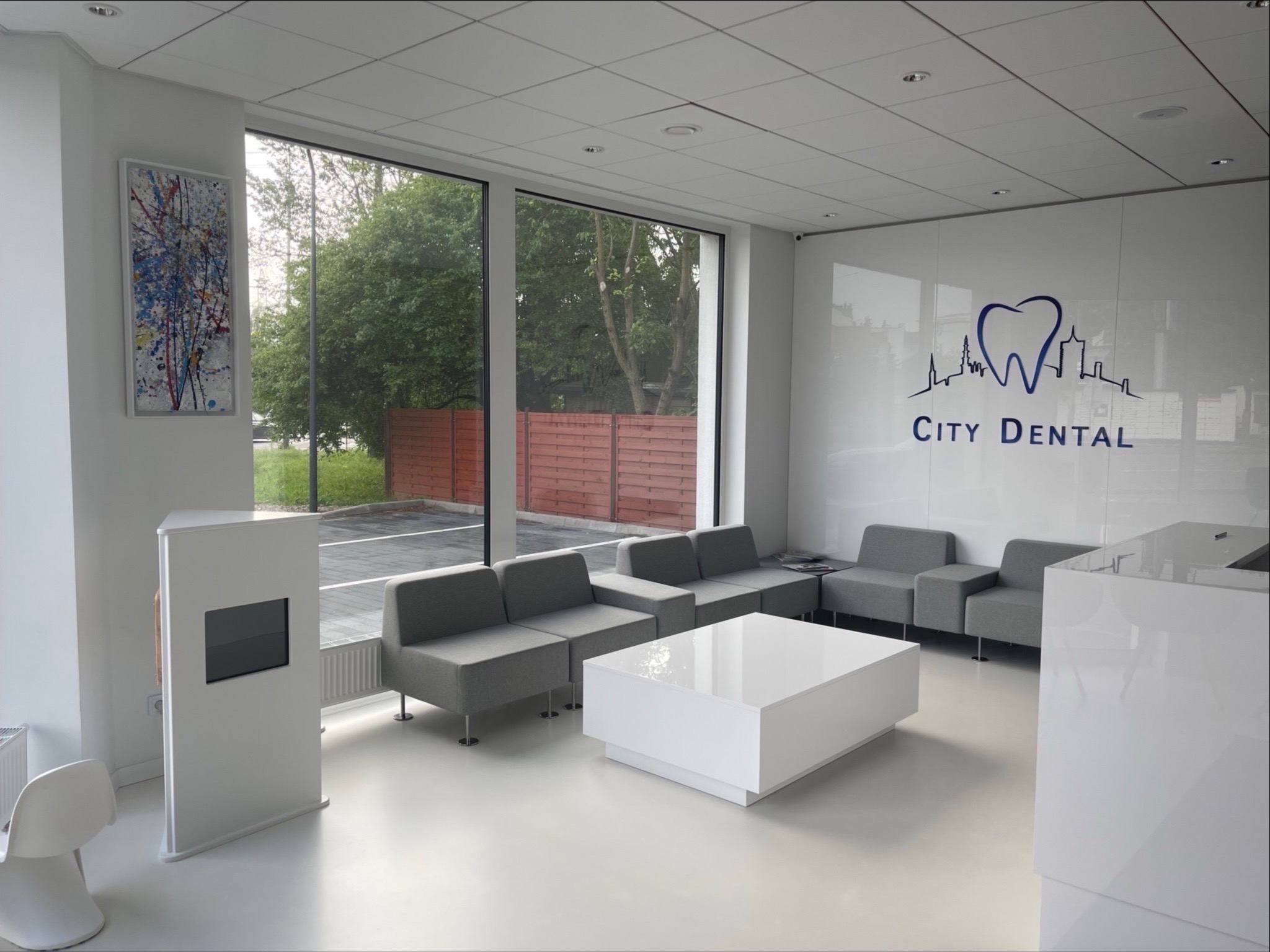 Jasne, minimalistyczne wnętrze poczekalni kliniki z szarymi sofami, białym stolikiem i recepcją, widoczne logo 'City Dental' na ścianie, obraz abstrakcyjny na ścianie po lewej stronie, duże okna...