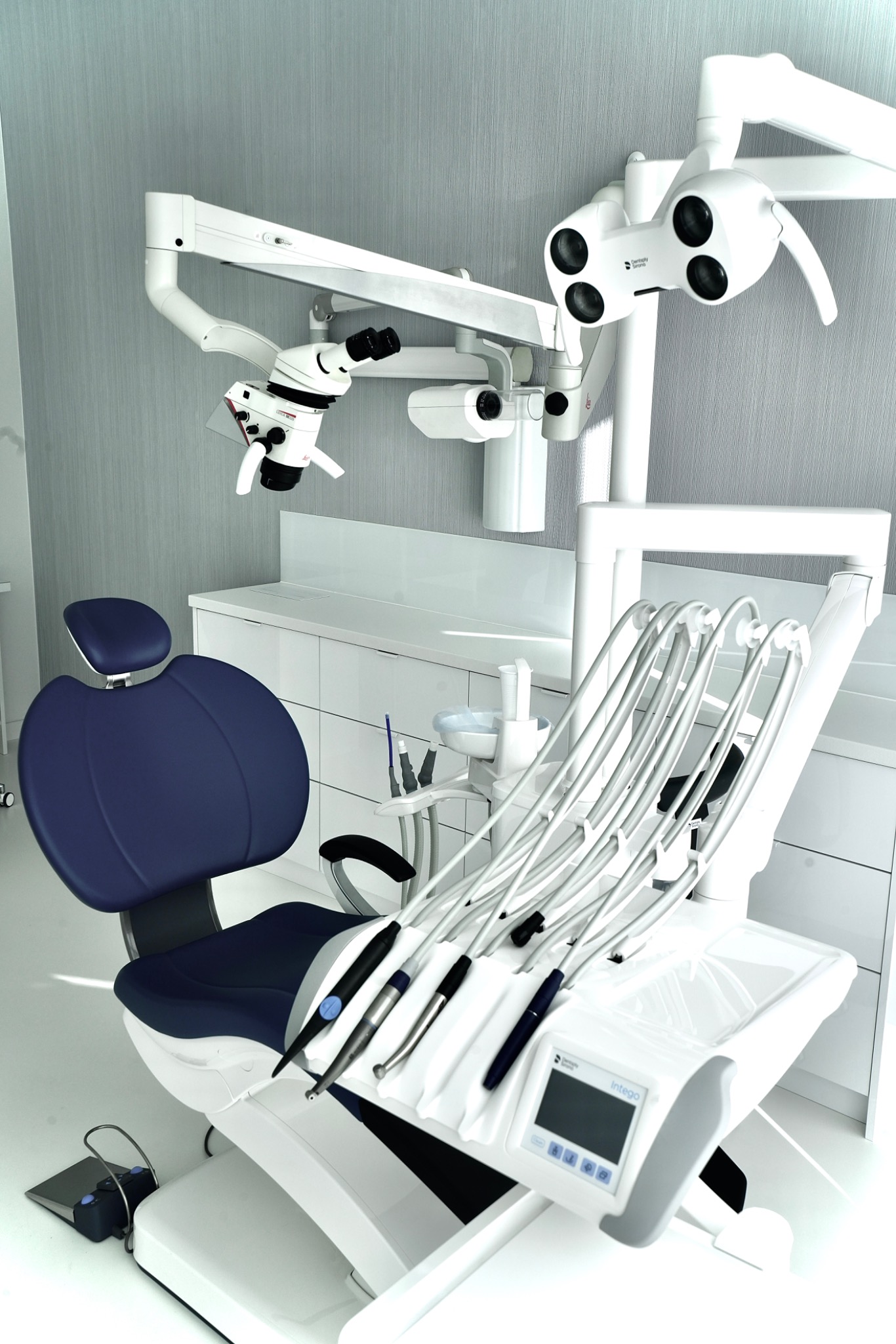 Nowoczesny unit stomatologiczny z niebieskim fotelem, oświetleniem i mikroskopem zabiegowym w minimalistycznym gabinecie.