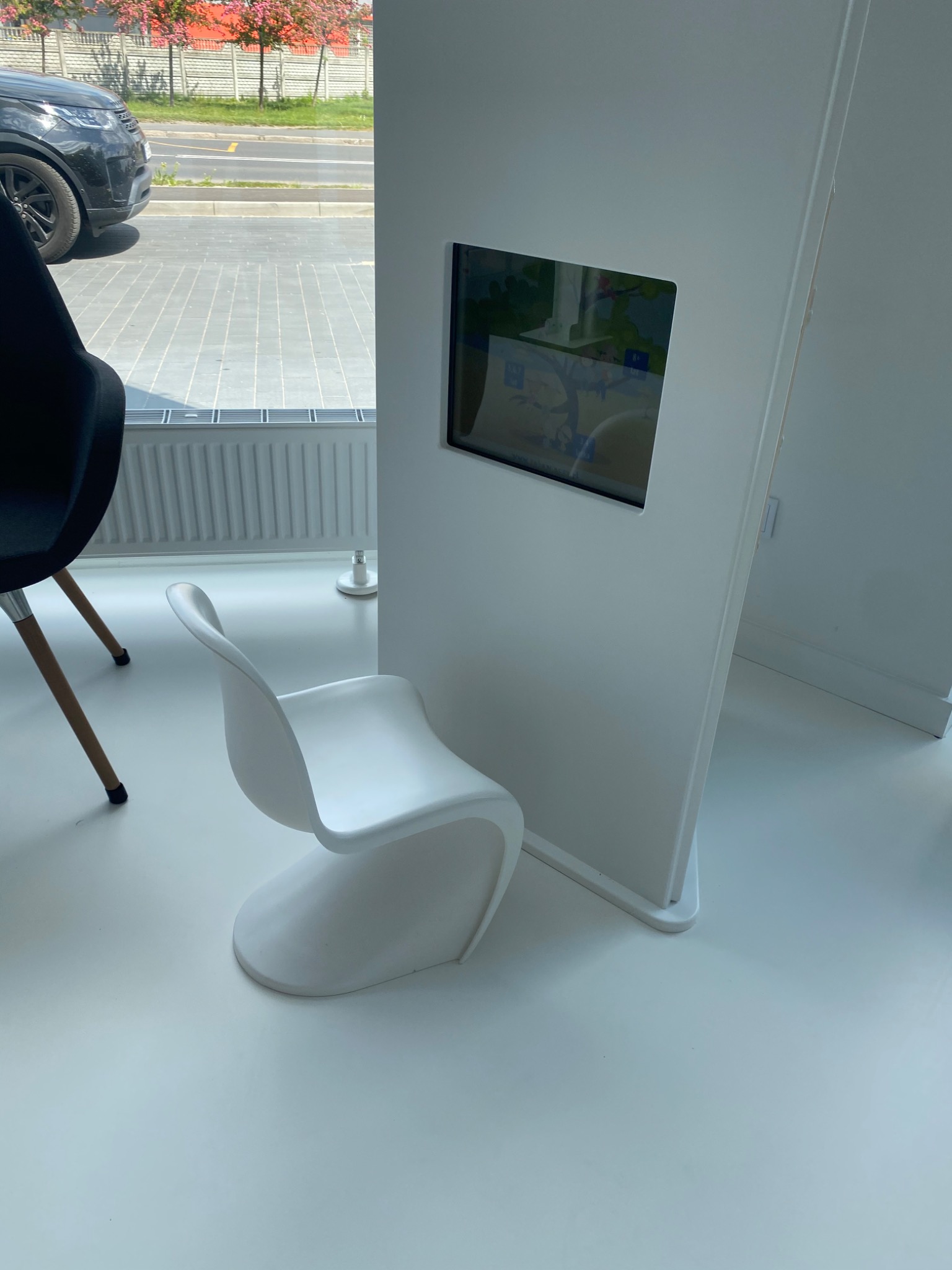Wnętrze poczekalni z białą podłogą, designerskim krzesłem i panelem z wbudowanym ekranem prezentującym mapę.