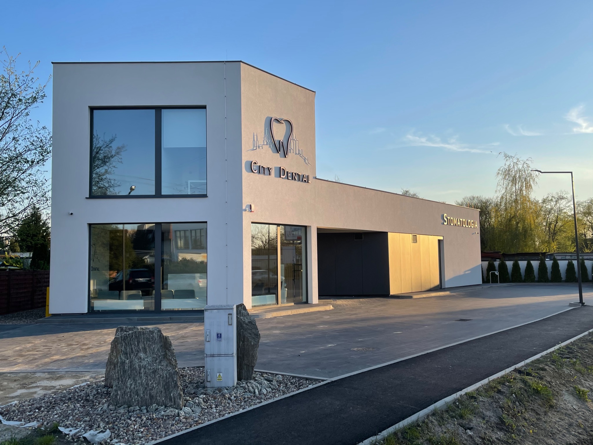 Nowoczesny budynek kliniki dentystycznej z dużymi oknami i logo w kształcie zęba na tle panoramy miasta, oświetlony popołudniowym słońcem, z kamieniami dekoracyjnymi przed wejściem.