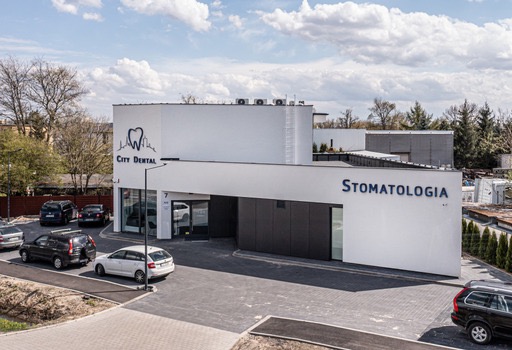 Nowoczesny budynek kliniki stomatologicznej City Dental z logo przedstawiającym ząb na tle panoramy miasta, widok z góry na parking przed budynkiem z zaparkowanymi samochodami.