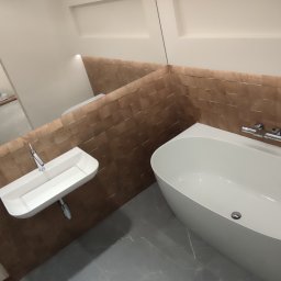 Mixa-Bud - Nowoczesna łazienka w trakcie prac wykończeniowych: biała wanna i umywalka na tle ściany wyłożonej drewnianymi panelami, widoczne odbicie osoby robiącej zdjęcie w lustrze.