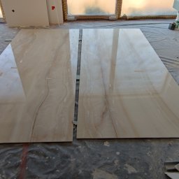 Mixa-Bud - Dwie duże, błyszczące płytki imitujące marmur leżą na podłodze zabezpieczonej folią, widoczne profile metalowe i ślady prac remontowych.