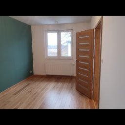 Mixa-Bud - Pusty pokój po remoncie z drewnianą podłogą, ścianą pomalowaną na ciemnozielono, białą ścianą, grzejnikiem pod oknem i nowoczesnymi drewnianymi drzwiami z przeszkleniami.