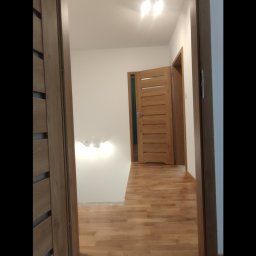 Mixa-Bud - Wnętrze mieszkania po remoncie: widok z korytarza na drewnianą podłogę, białe ściany z wystającymi kablami i otwarte drewniane drzwi.