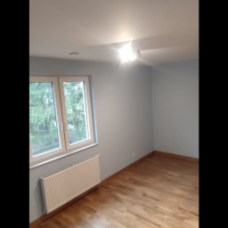 Mixa-Bud - Pusty pokój po remoncie z drewnianą podłogą, świeżo pomalowanymi ścianami na jasny niebieski kolor, białym grzejnikiem pod oknem i widokiem na drzewa za oknem.