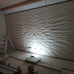 Mixa-Bud - Montaż dekoracyjnej, trójwymiarowej ściany z paneli gipsowych o falistym wzorze w trakcie prac wykończeniowych w mieszkaniu w Warszawie, widoczne narzędzia i drabina.