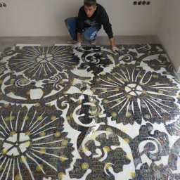 Mixa-Bud - Pracownik w rękawicach układa skomplikowaną mozaikę podłogową z motywem kwiatowym, zabezpieczoną folią ochronną, w surowym wnętrzu z widocznymi puszkami elektrycznymi.