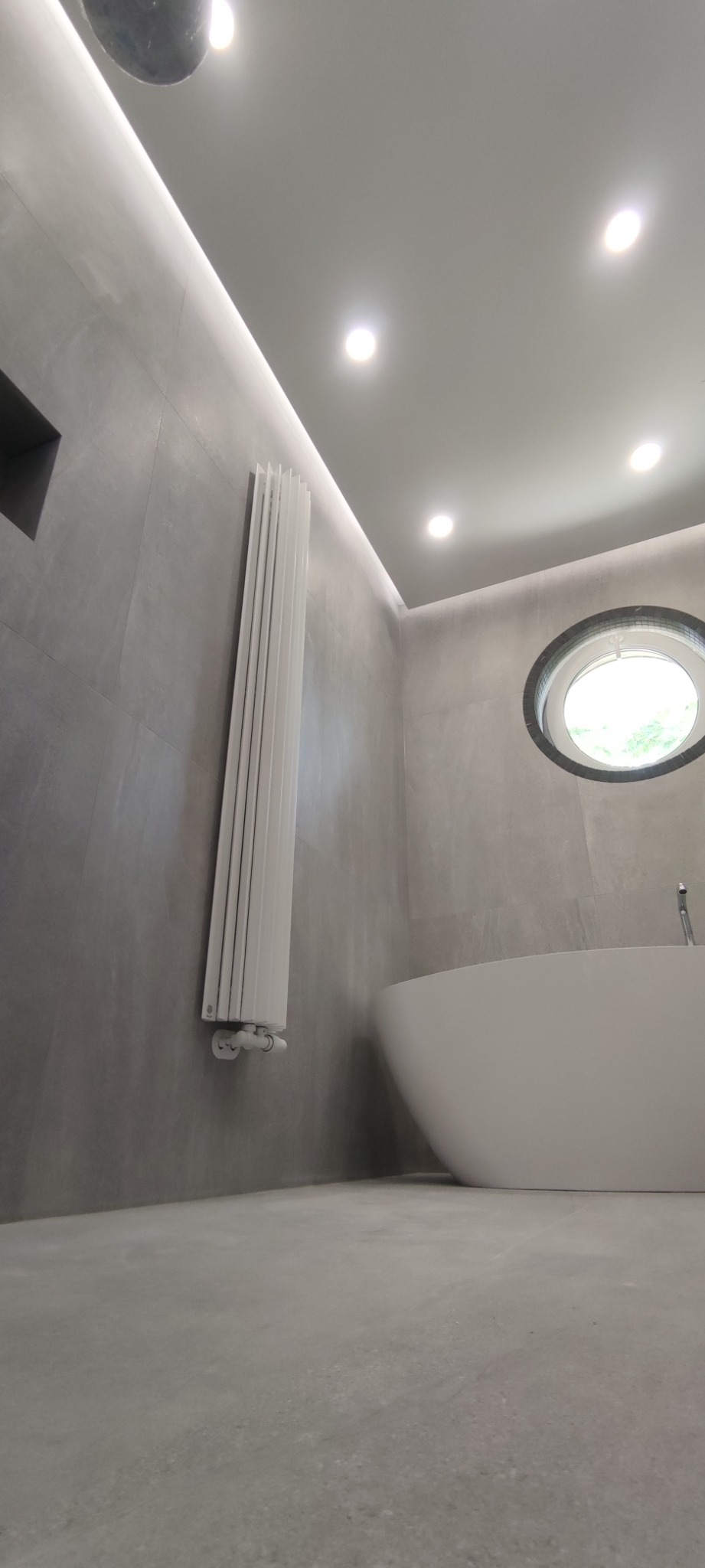 Minimalistyczna łazienka w odcieniach szarości z wolnostojącą białą wanną, okrągłym oknem i pionowym grzejnikiem żeberkowym, oświetlona punktowymi lampami sufitowymi i listwą LED.