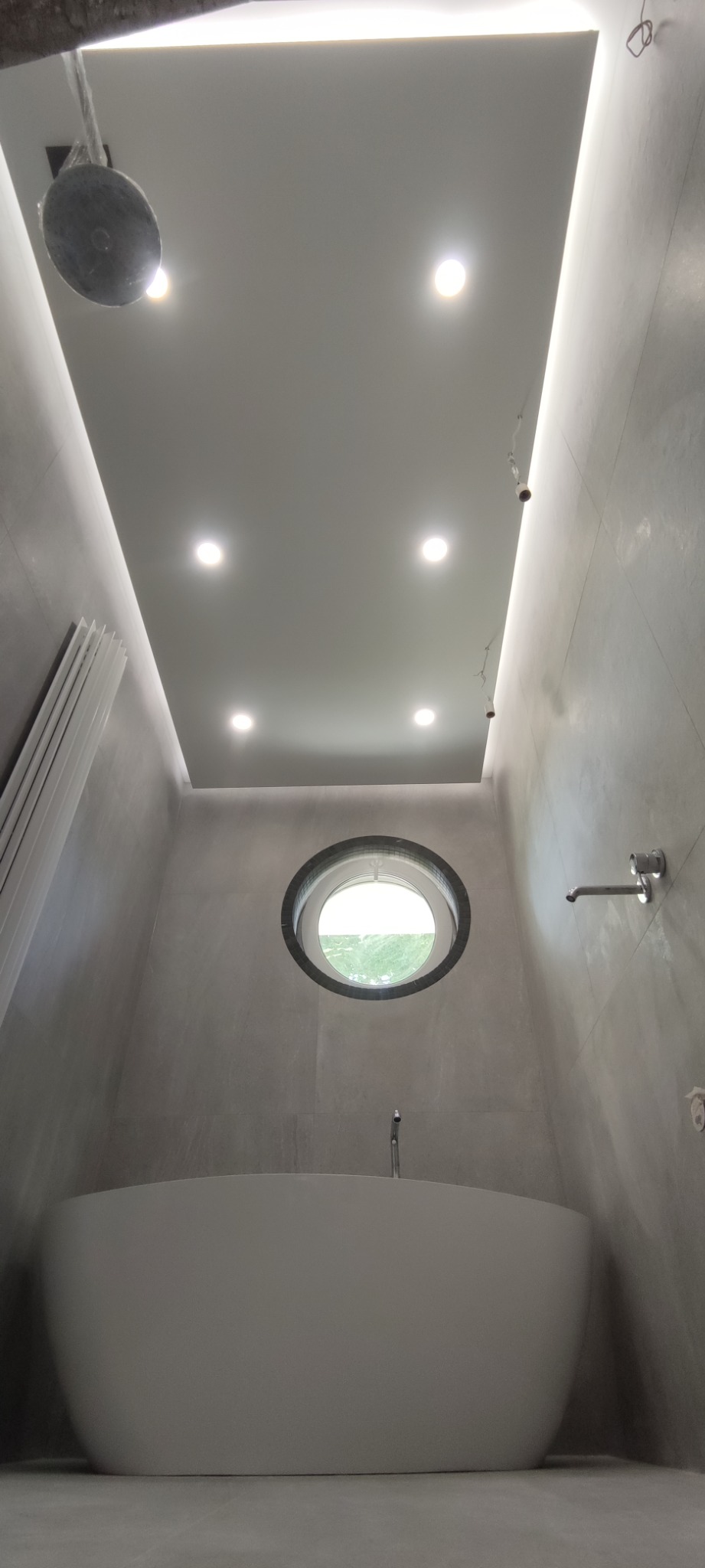 Nowoczesna łazienka w szarościach z wolnostojącą wanną, okrągłym oknem i oświetleniem LED w suficie podwieszanym.
